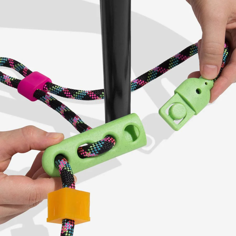 Hands adjust colourful rope accessories around a black pole, featuring green, pink, and orange components for securing and managing the rope.