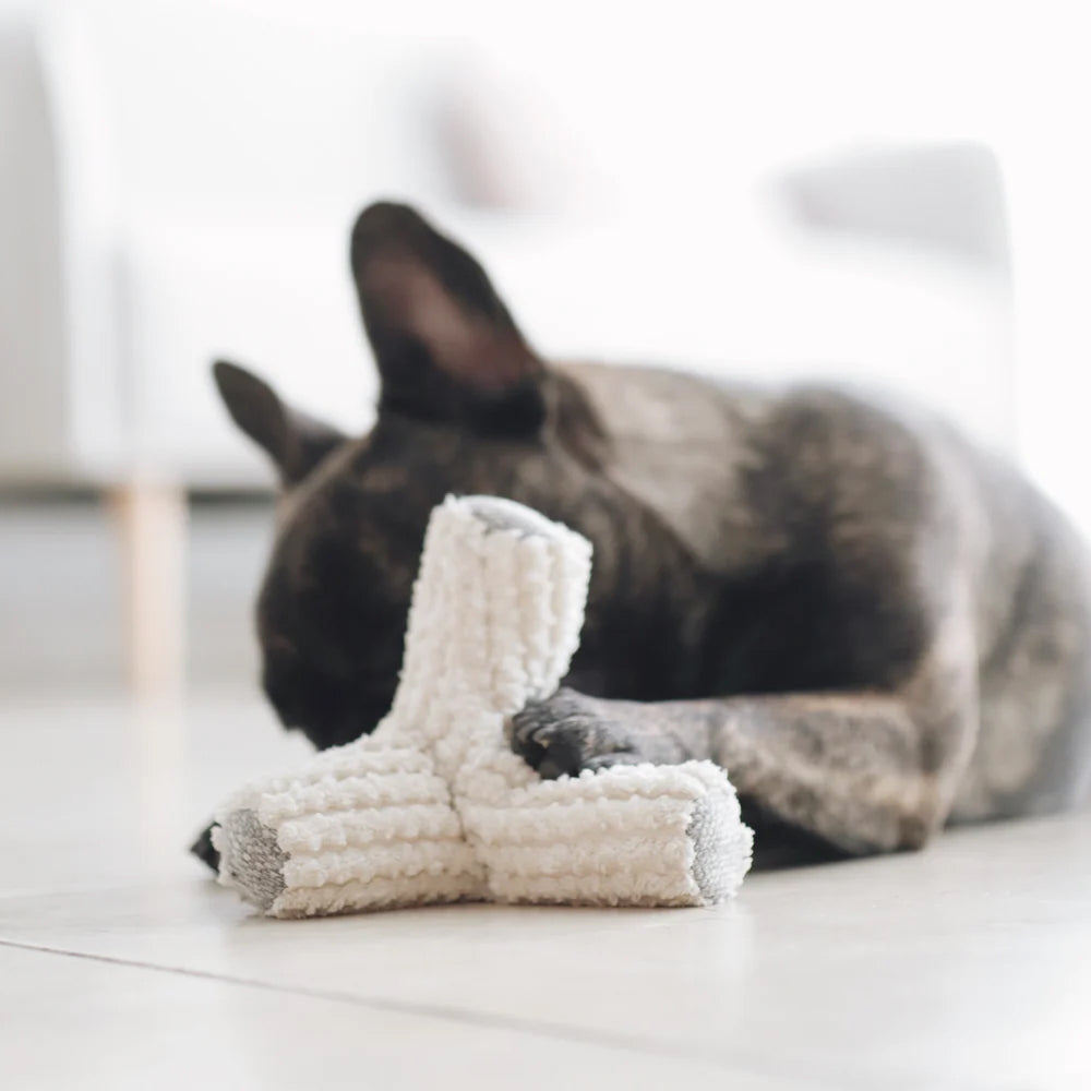 A relaxed brindle French Bulldog lays on the floor with a fluffy, white chew toy, softly resting its head on it.