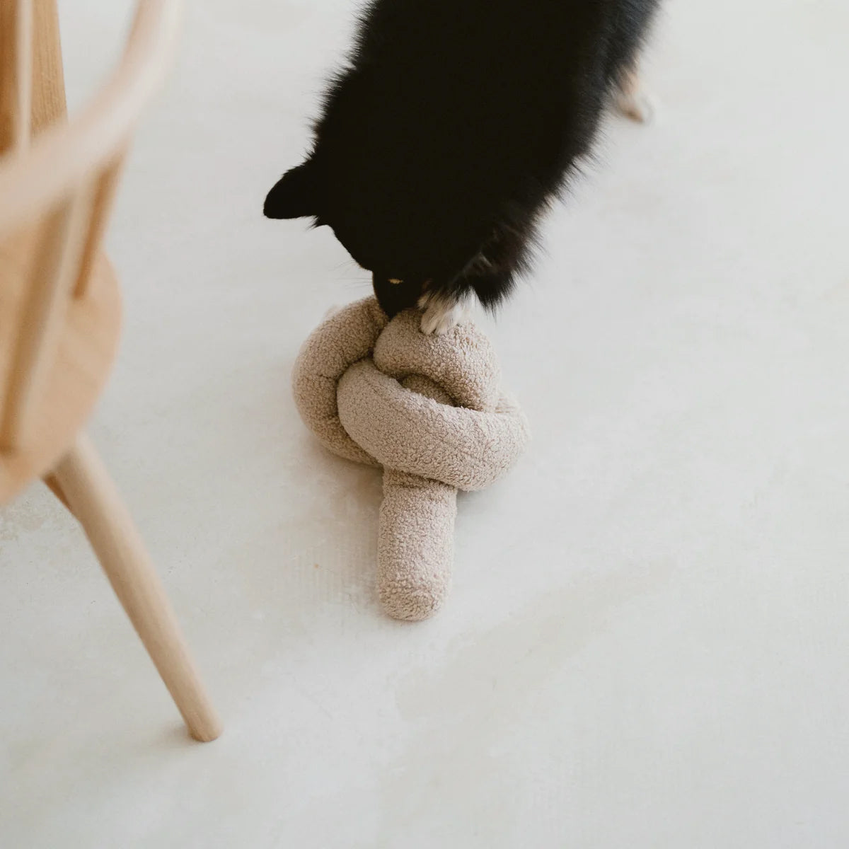 A black dog with a fluffy coat plays with a beige, knotted plush toy on a pale floor, showcasing a moment of playful interaction.