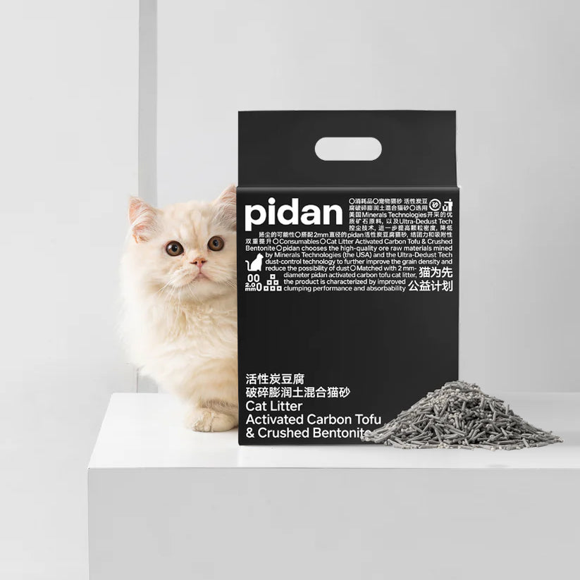 A fluffy white cat beside a black package of 'pidan' cat litter with text and a pile of litter.