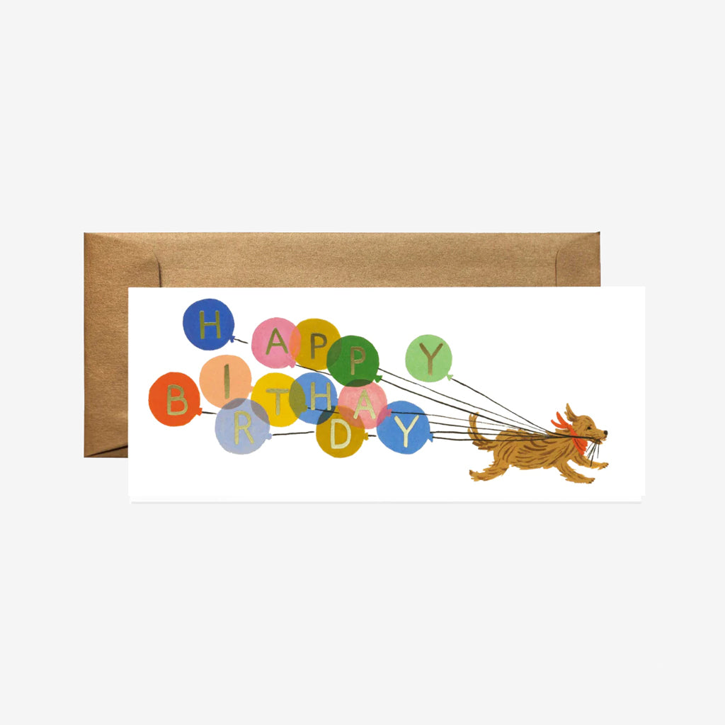 A playful dog pulls colourful balloons with the phrase "Happy Birthday" in a whimsical design on a card next to a brown envelope.