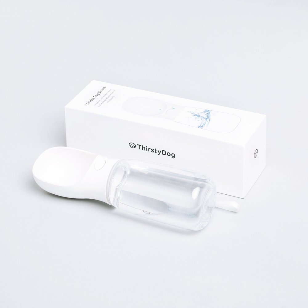 A white Thirsty Dog portable water bottle with a removable bowl, positioned next to its packaging on a light gray background.