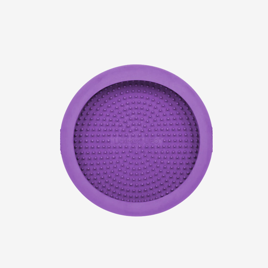 A round, textured, purple silicone mat with raised dots, designed for pet grooming or feeding, featuring a central logo.
