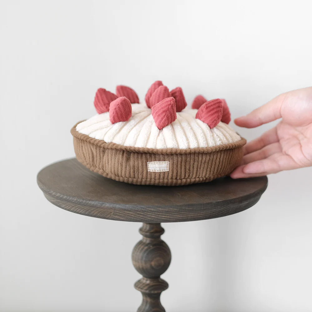 A plush dessert toy resembling a pie, featuring a creamy top and pink fabric toppings, placed on a wooden table.