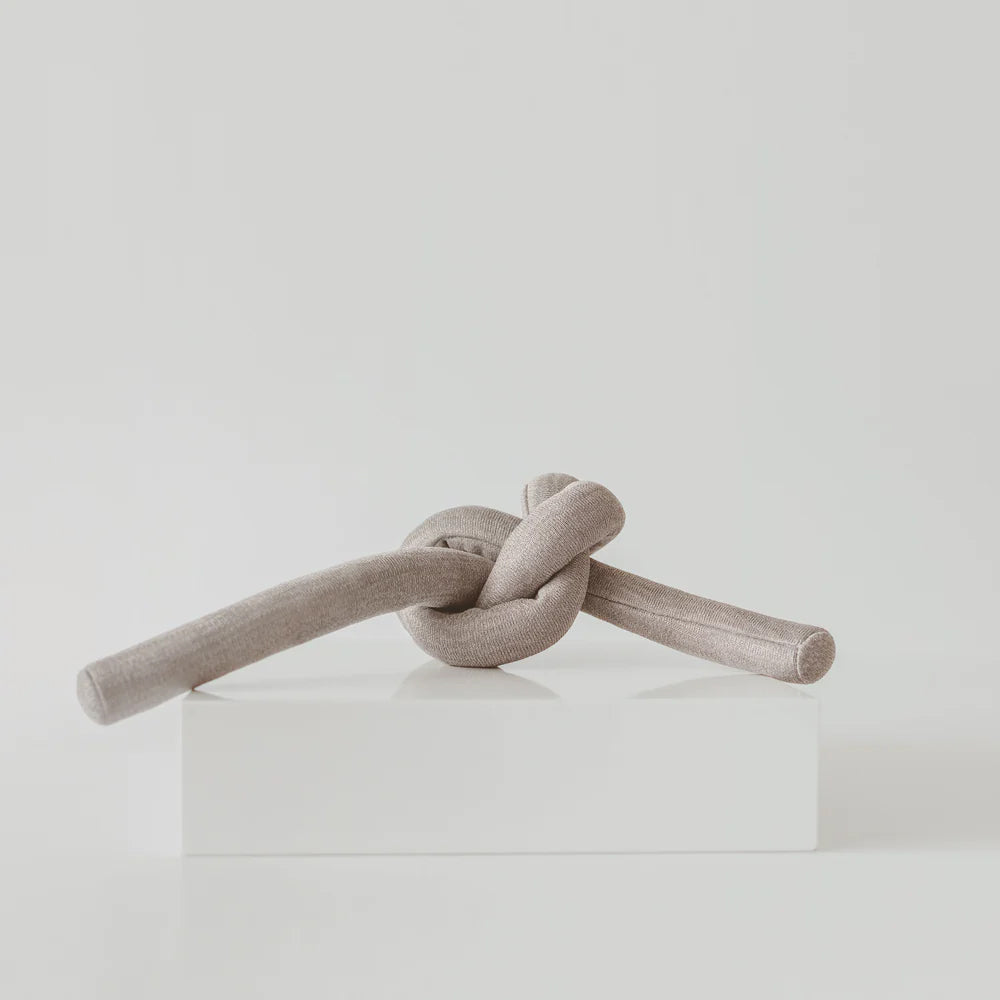 A soft, beige fabric knot sculpture resting on a white pedestal against a minimalistic background.