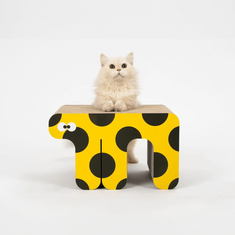 A fluffy white cat sitting on a yellow and black polka-dot cartoon elephant cat scratcher.