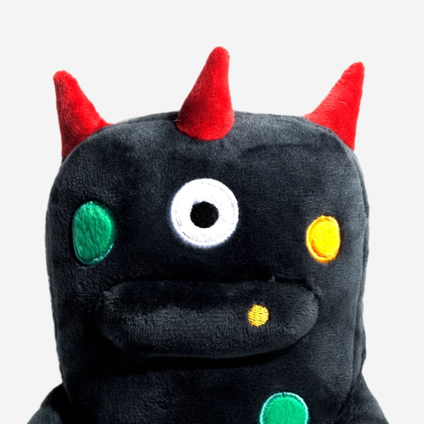 A navy blue plush alien toy with red horns, one eye, and colourful spots on a white background.