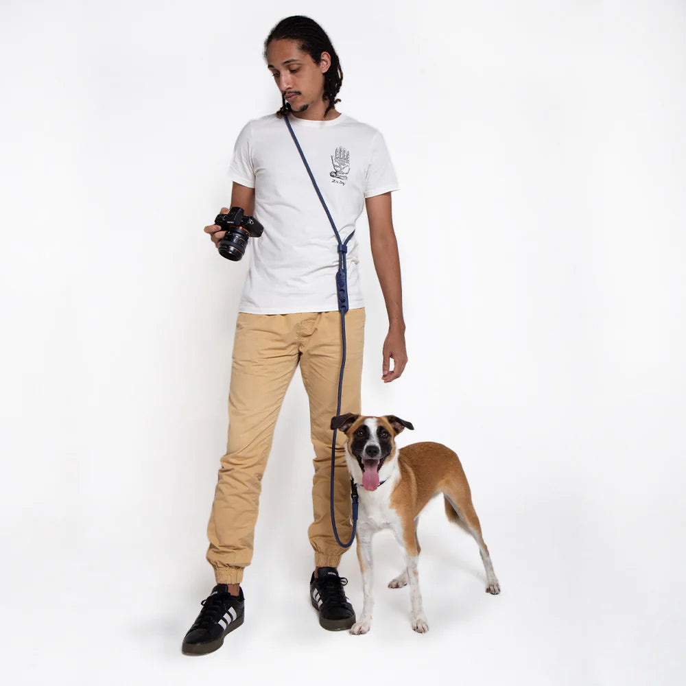 A person holding a camera stands next to a happy dog on a leash, dressed in a white t-shirt and khaki pants against a plain background.