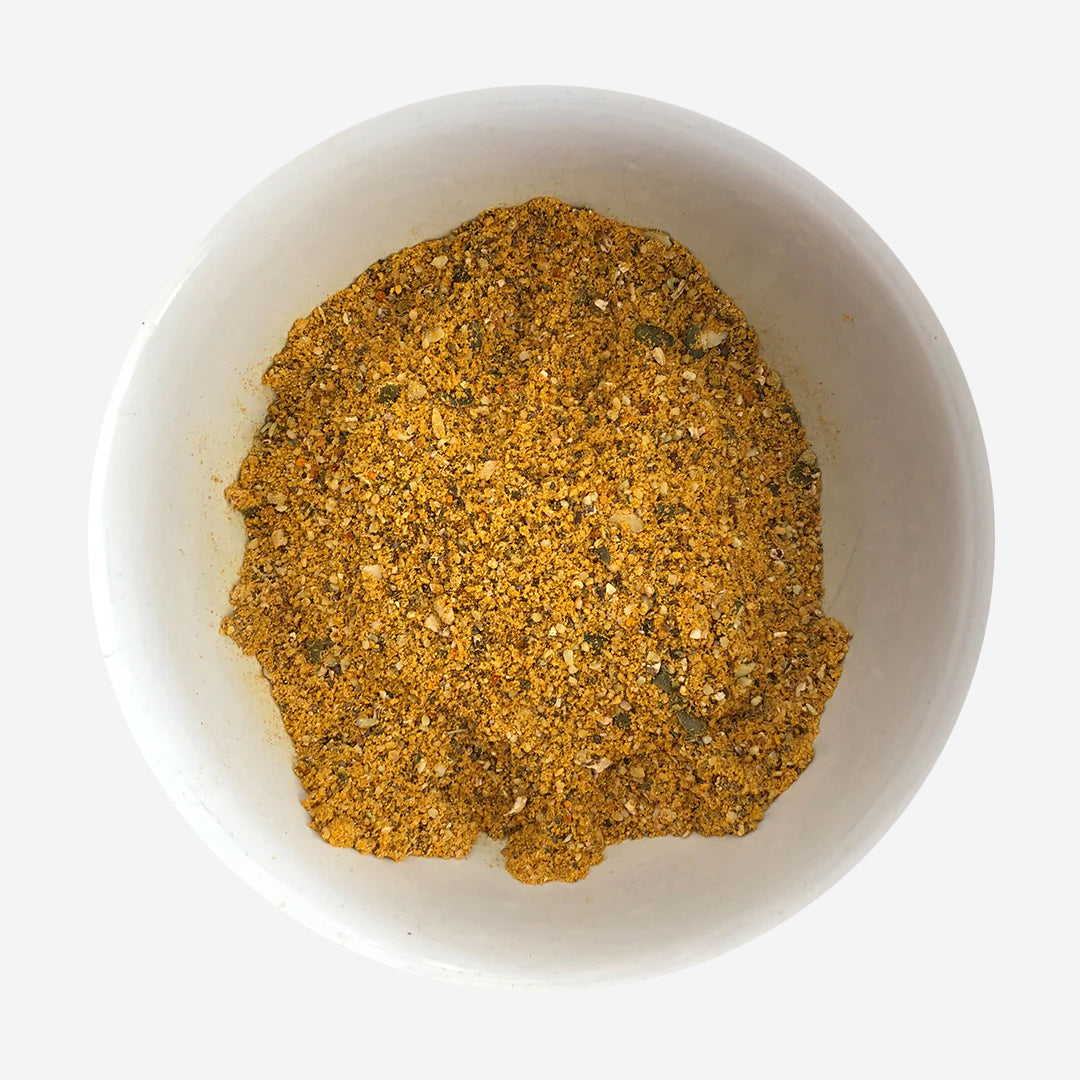 A close-up of a white bowl filled with a coarse, golden-yellow spice blend, featuring a mix of herbs and textured grains.