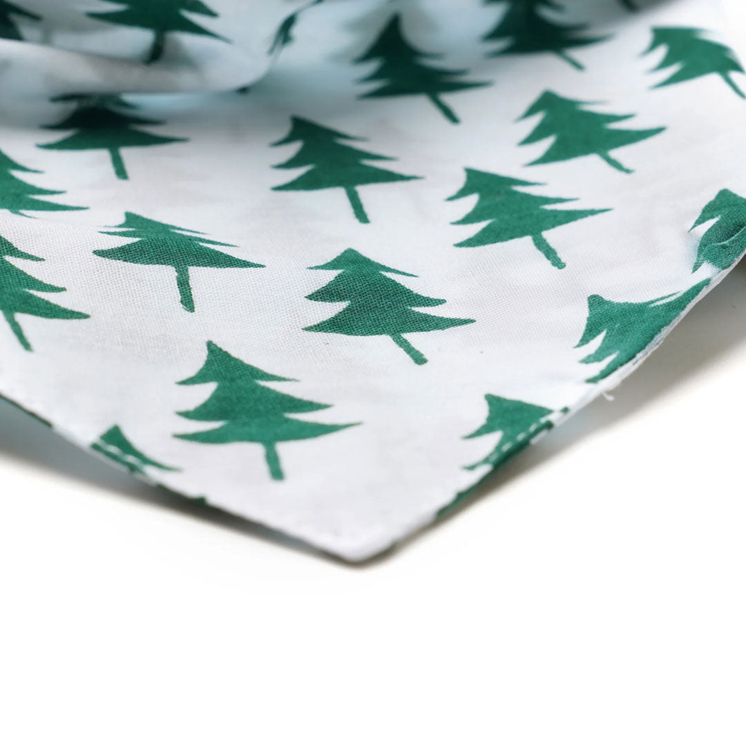 Close up fabric of a white dog bandana with a nature-inspired design featuring green Christmas trees printed on it.