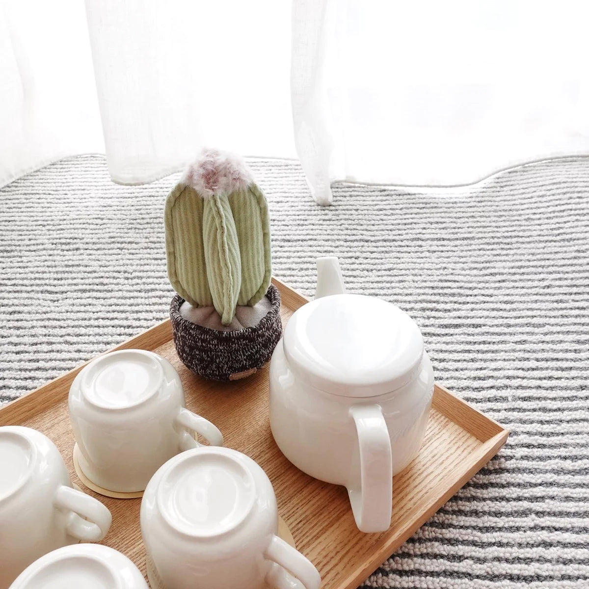 White teapot and cups sat on a bamboo tray, with a A plush pale green cactus dog toy, with pink fluff at the top sat in a soft brown shaped pot, in front of a white curtain.