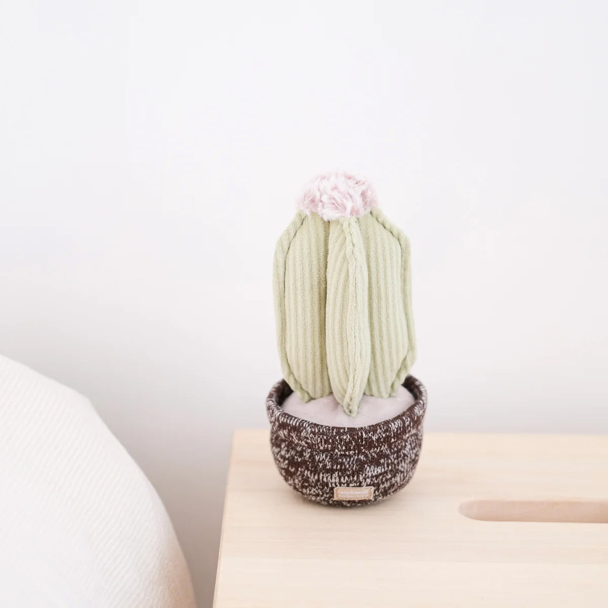 A plush pale green cactus dog toy, with pink fluff at the top sat in a soft brown shaped pot. Sat on a minimal table top.