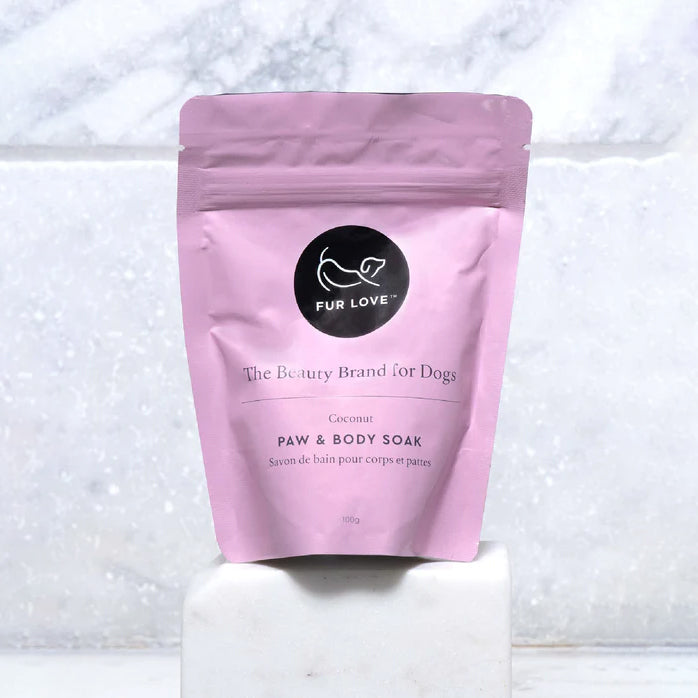 A pink pouch filled with paw and body soak, sat in front of a marble background.