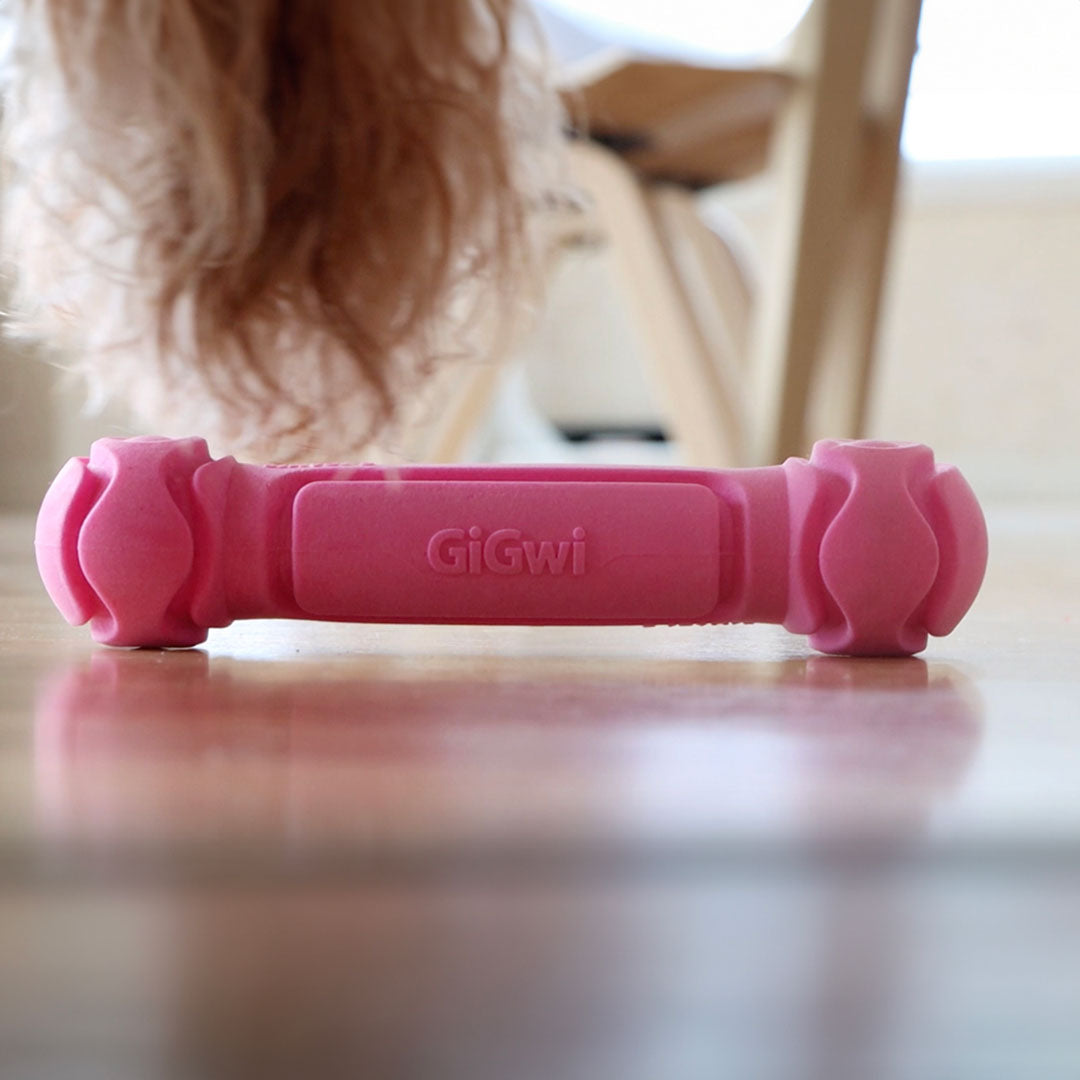 A pink foam and rubber dumbbell dog toy sat on a wooden floor. With a floofy dogs ear in the foreground.