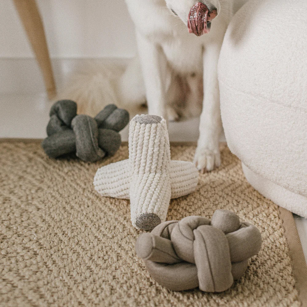 Three plush dog toys sitting on a stone coloured rug, with a white dog in the background.