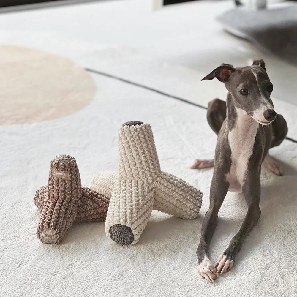 A small grey whippet lying on a pale grey carpet with 2 plush dog toys.