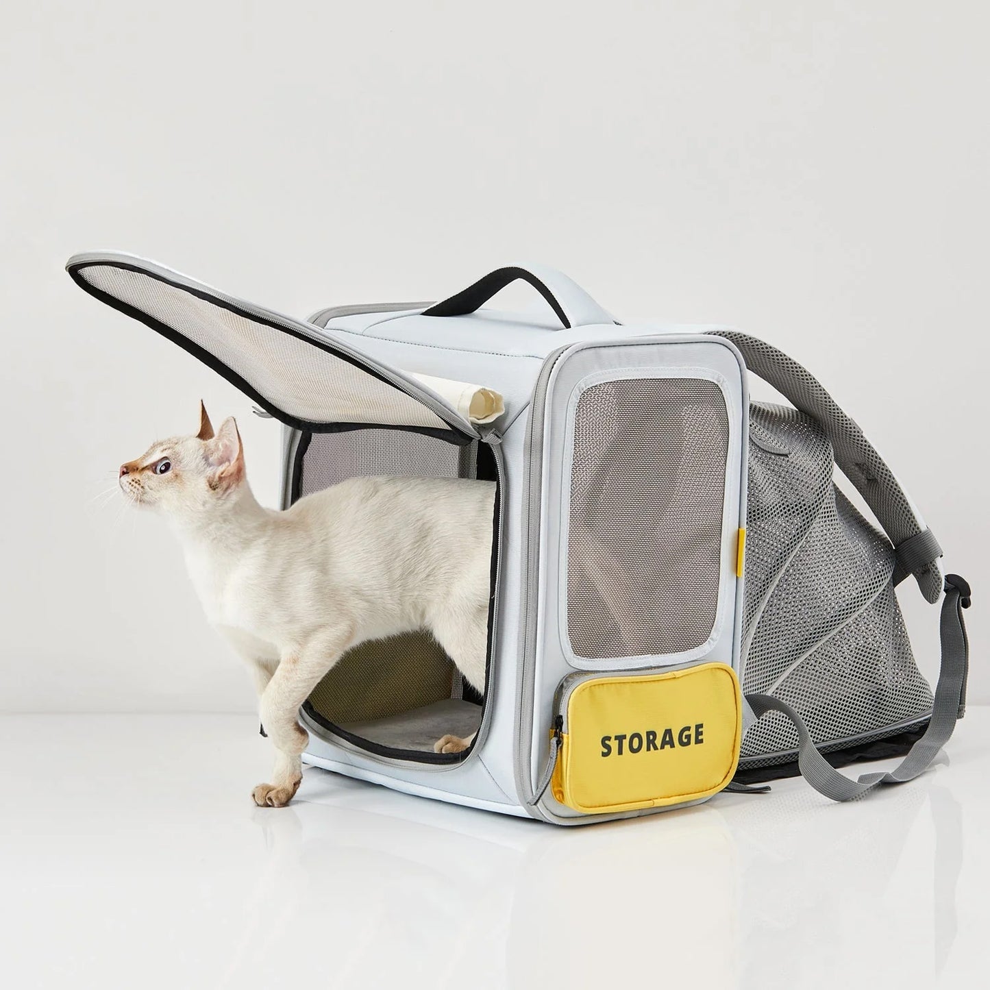 A white cat stepping out from a grey pet backpack by PetKit with extra tent space, featuring a yellow storage compartment