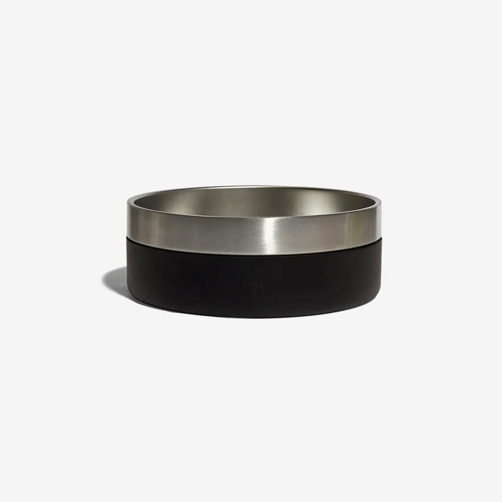 A stainless steel dog bowl with a black rubber exterior by Zee Dog with kibble in resting against a plain white background.