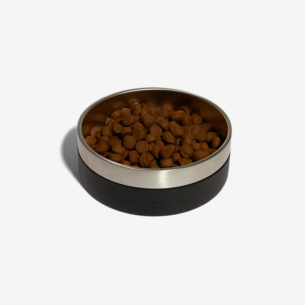 A stainless steel dog bowl with a black rubber exterior by Zee Dog with kibble in resting against a plain white background.