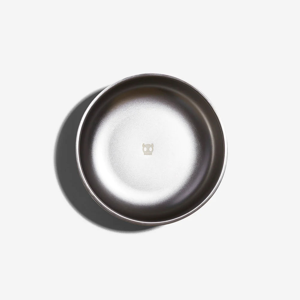 A stainless steel dog bowl with a black rubber exterior by Zee Dog with their skull icon embossed in the center, viewed from above on a white background.