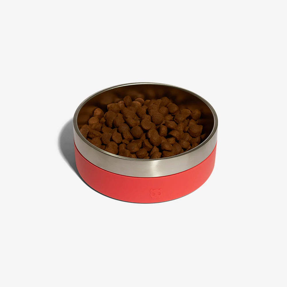 A stainless steel dog bowl with a red rubber exterior by Zee Dog with kibble in resting against a plain white background.