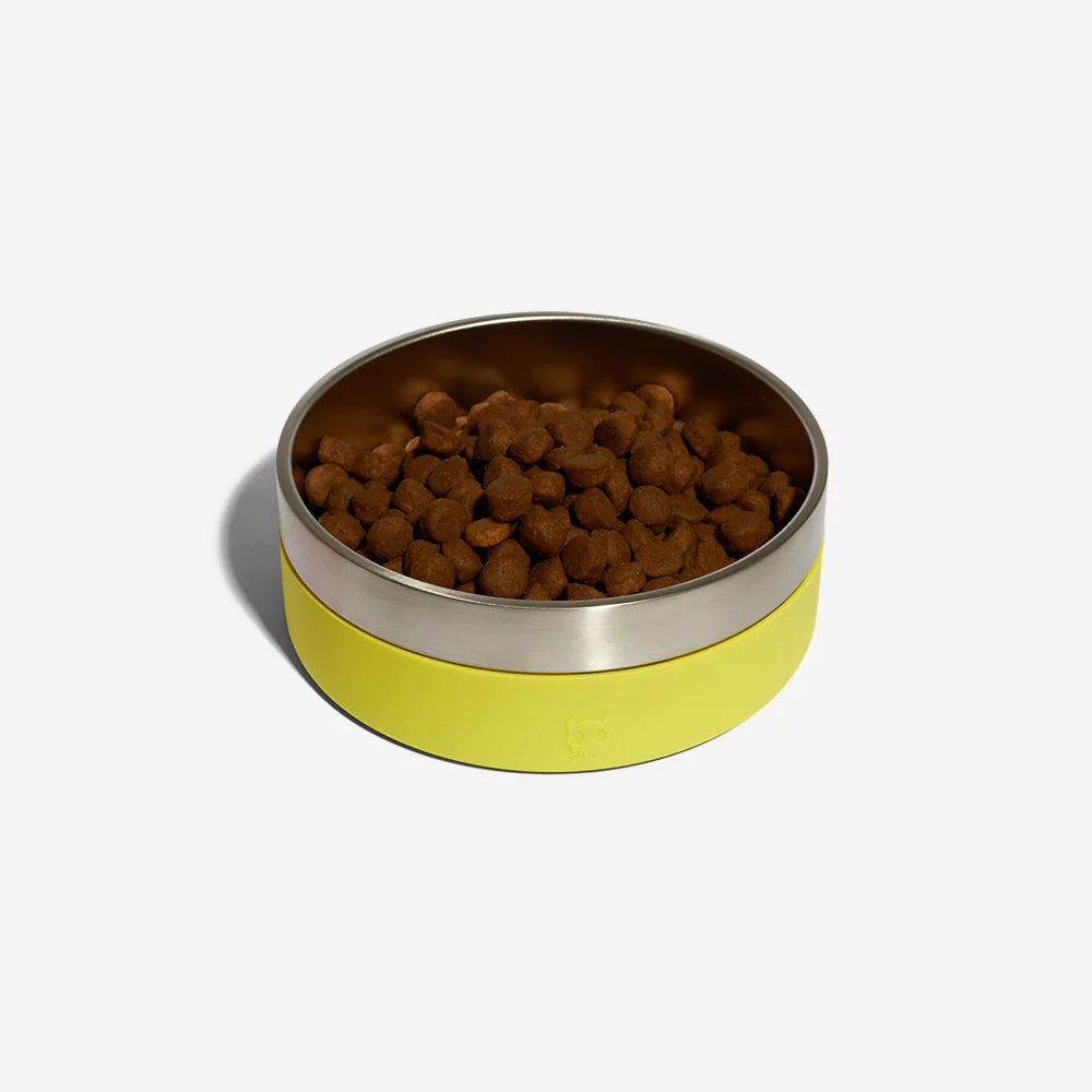 A stainless steel dog bowl with a black rubber exterior by Zee Dog with kibble in resting against a plain white background.